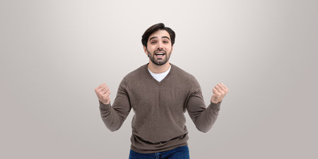 Overjoyed young man with beard and fists up, happy winner on empty copy space beige background. Concept of celebration of success, profit, lottery and luckの写真素材