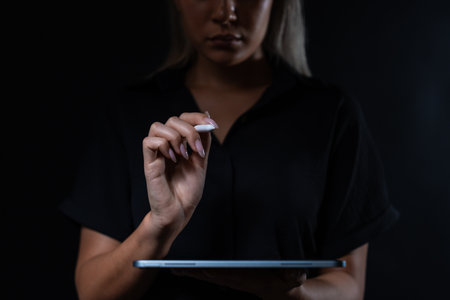 Woman's hand holding a stylus, interacting with a tablet in a dark setting. Concept of technology and digital creativityの写真素材