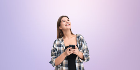 Smiling woman holding a smartphone, wearing a plaid shirt, looking up on a light purple background. Concept of social media and online networkingの写真素材