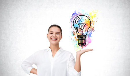 Woman smiling confidently in white shirt, holding a vibrant, colorful abstract light bulb on white background, symbolizing creativityの写真素材
