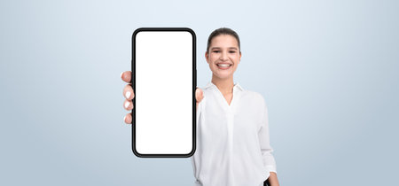Smiling woman holding a smartphone with a blank screen, dressed in a white shirt, on a light background. Concept of mobile technology and businessの写真素材