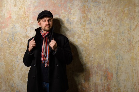 image of a caucasian man with grungy blond hair dressed warmly in a stylish black trench coat and a funky hat and scarf for the cold winterの写真素材