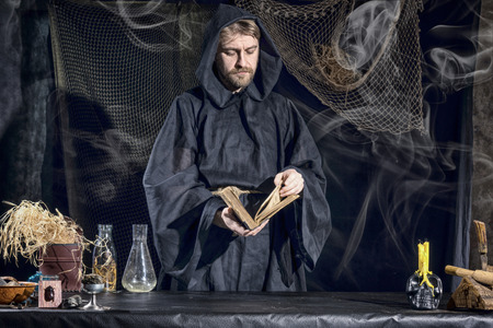 The medieval alchemist reading a book at the table in his laboratory smoke in the background....