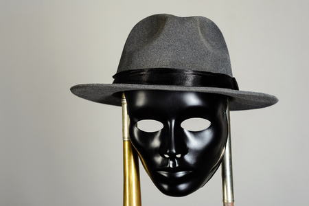 Black theater mask and hat hanging on a brass pipe on gray background with copy space. Social masking and mystery conceptの写真素材