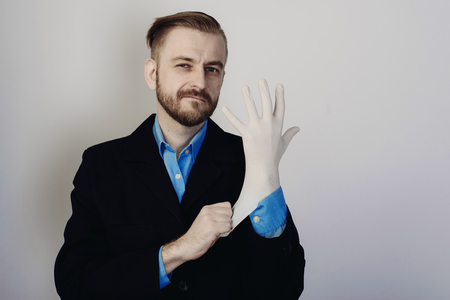 young stylish bearded businessman in suit and blue shirt puts on medical glove on gray backgroundの写真素材