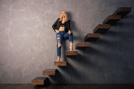 Beautiful woman in a black leather jacket and torn jeans posing standing on a wooden cantilever staircase.の写真素材