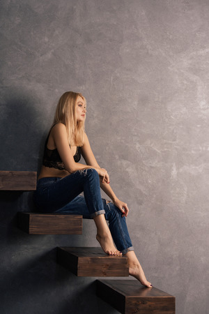 beautiful sensual woman in a bra and jeans sets on a wooden cantilever steps. Looking awayの写真素材