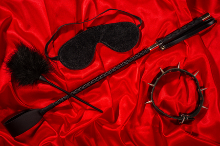 Bondage, kinky adult sex games, kink and BDSM lifestyle concept with awhip, feather stick, collar, eye mask on red silk with copy spaceの写真素材