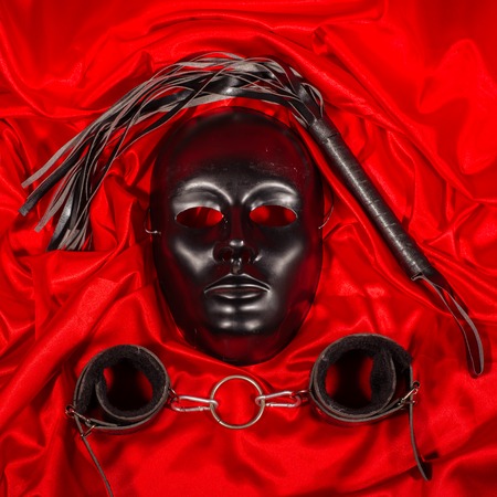 Bondage, kinky adult sex games, kink and BDSM lifestyle concept with a mask, pair of leather handcuffs, flogger, ball gag and a coller with a leash attached on red silk with copy spaceの写真素材