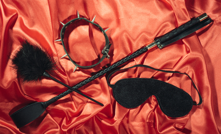 Bondage, kinky adult sex games, kink and BDSM lifestyle concept with a whip, feather stick, collar, eye mask on red silk with copy spaceの写真素材