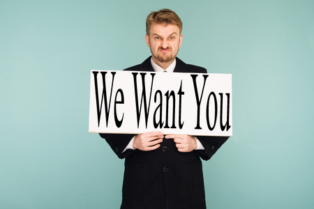 Angry beard young business man showing signboard with sign We Want You, on blue backgroundの写真素材