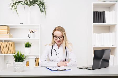 Young beautiful doctor woman working happy and smile in hospital, sitting on table, medical conceptの写真素材