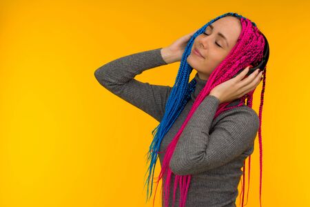 Happy cheerful woman with senegalese braids and freckles wearing wireless headphones listening to music from smartphone studio shot isolated on yellow backgroundの写真素材