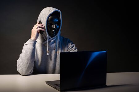 Anonymous hacker in mask programmer uses a laptop and talking on the phone to hack the system in the...