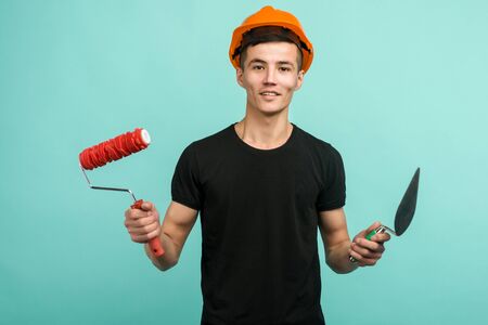 Asian working man in an orange helmet stands with a paint roller and trowel on a blue background -...
