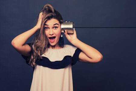 portrait of young surprised woman hearing through a tin can over grey.の写真素材