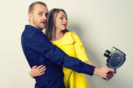 Surprised man in a blue shirt and a young slim girl in a yellow dress are hugging and shooting themselves on an old movie cameraの写真素材