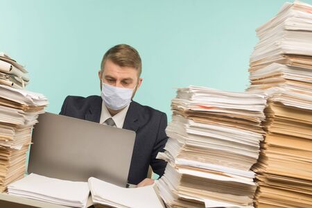 A male accountant or company manager works in an office in a pandemic in view of the accumulated paper work. A protective medical mask is on the face. On the desktop are large stacks of documents.の写真素材
