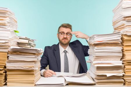 Shocked businessman sitting at the table with many papers in office, he is overloaded with work -...