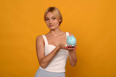 Beautiful young girl holding a pig piggy bank on yellow background. For saving money wealth and...