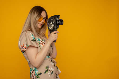 Beauty with movie camera. Cheerful young woman holding movie camera and posing against yellow...