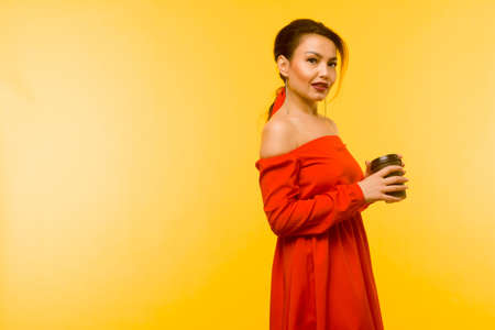 Beautiful woman in red dress posing with coffee cup on yellow backgroundの写真素材