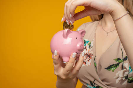 Young positive woman holding a pink piggy bank and bitcoin in her hand on a yellow backgroundの写真素材