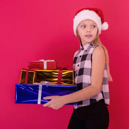 Funny smiling joyful child girl in Santa hat holding Christmas gift in hand over the red background - imageの写真素材