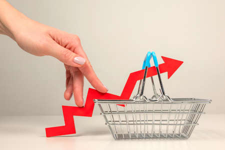 The fingers of a woman's hand step up the arrow of a chart lying on a shopping basket. Crisis and rising commodity prices conceptの写真素材