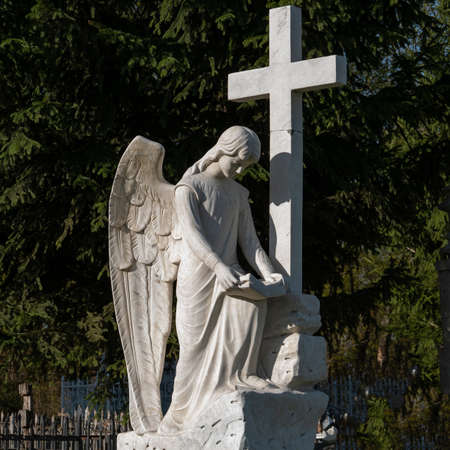 Beautiful sad angel with cross. Fragment of ancient stone statue with a sweet expression that looks down as a symbol of unspeakable sadness and death.の写真素材