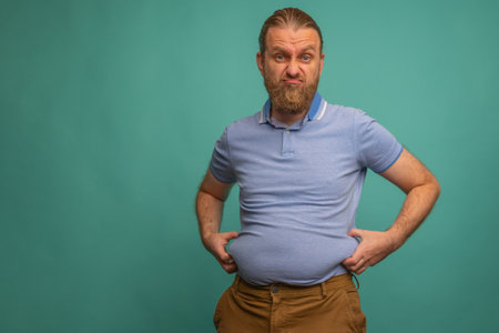 A man with a sad face feels his fat belly on blue background, closeup. Space for textの写真素材
