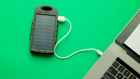 A modern laptop is being charged from a solar-powered charging station on a green background. The...