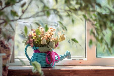 Decorative vase - a blue wicker watering can with a pink bow with dry wildflowers herbarium, stands on the windowsill of a country house in the village . Design interior, rustic style. countryside lifeの写真素材