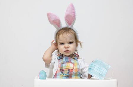 Angry toddler child kid with easter bunny ears holds a medical mask. Easter and quarantine concept...