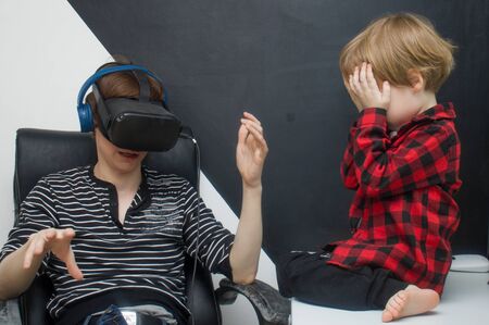 A young father plays in a virtual reality helmet. Little boy son is sitting nearby and crying. Child...