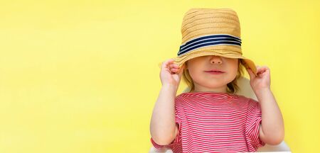 Little toddler girl hides her face with a straw hat. The child pulled a hat on his head on a yellow background. Copy space and place for text.の写真素材