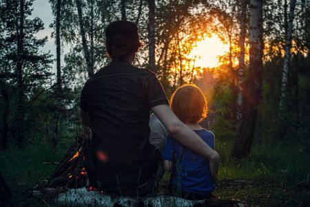 Father and son are sitting sitting on a log by the fire and watching the sunset with their backs. Forest background.の写真素材