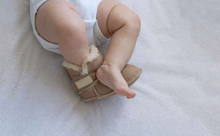 Feet of a newborn toddler close-up on the bed. On one foot boots made of leather and fur. The second...