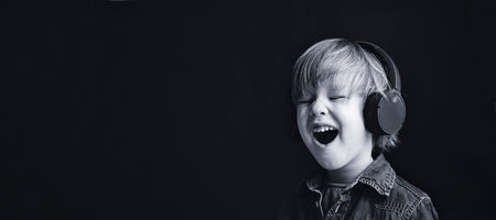 Little child boy with closed eyes listens with pleasure in headphones on a black background with a place for text copy space banner.の写真素材