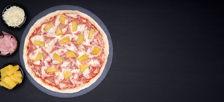 Hawaiian pizza made with pineapple, ham and mozzarella cheese on a stone plate with a black background with copy space for advertising. The pizza is accompanied by its main ingredients. An Italian pizza made with the best ingredients.の写真素材