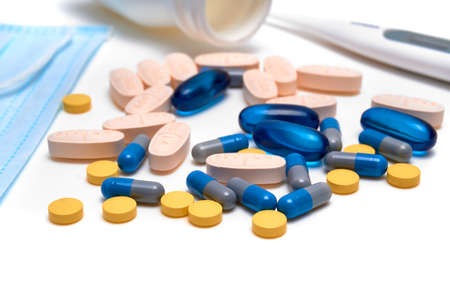 variety of capsules and pills with a bottle of pills, a thermometer and a face mask on a white...