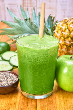 green smoothie of spinach, cucumber, lemon, pineapple and bamboo straw. fresh fruit on the side and a wooden bowl with chia seeds. wooden table with wooden planks and white wooden background.の写真素材