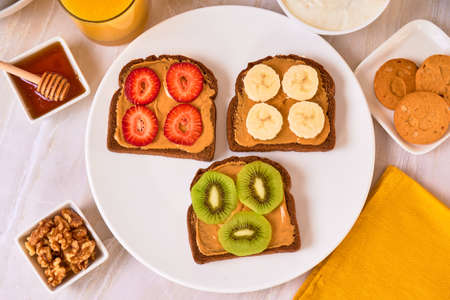 healthy low-calorie breakfast, toasted bread with peanut butter, strawberries, kiwi and banana, accompanied by fresh fruit and orange juice, on a marble surface and white background. top viewの写真素材