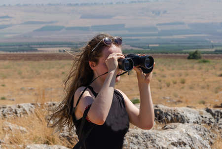 girl in a black T-shirt looks through binoculars into the distance in the mountainsの写真素材