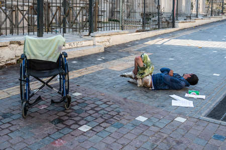 ATHENS, GREECE - MAY 14, 2022: A homeless man sleeps on a sidewalk in downtown Athens. Tourists pass by.の写真素材