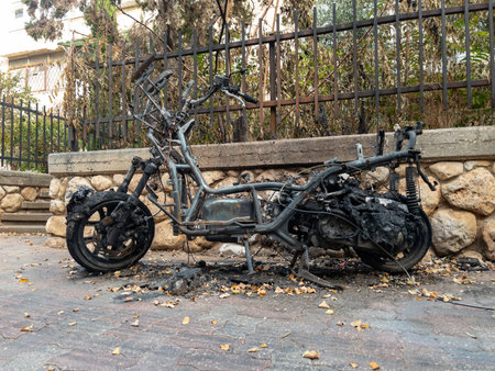 Burnt motorcycle frame after a fire on a city street. Charred metal, melted parts, and signs of destruction near a stone wall and iron fence. High quality photoの写真素材