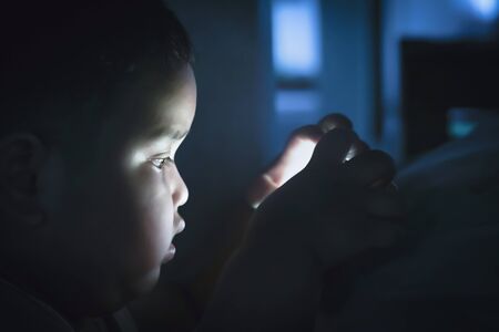 Fat boy playing smartphone in bedroom at night time on dark background. Prolonged telephone play...