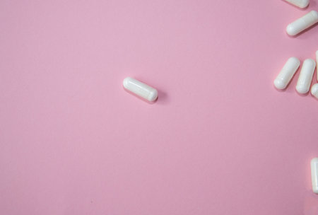 White capsules are scattered across a soft pink background, emphasizing the pharmaceutical product's...