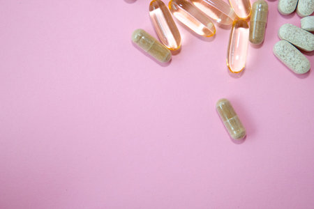 Various dietary supplements including capsules and softgels are scattered on a pink background,...