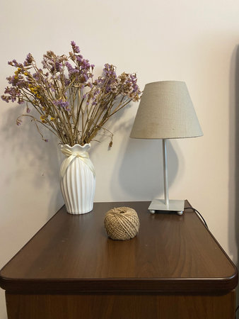 A white vase holds dried flowers beside a beige lamp on a wooden nightstand, with a natural twine ball adding texture to the arrangementの写真素材